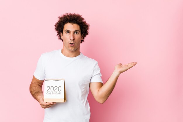 Jeune homme bouclé caucasien tenant un calendrier 2020 impressionné tenant copie espace sur la paume.