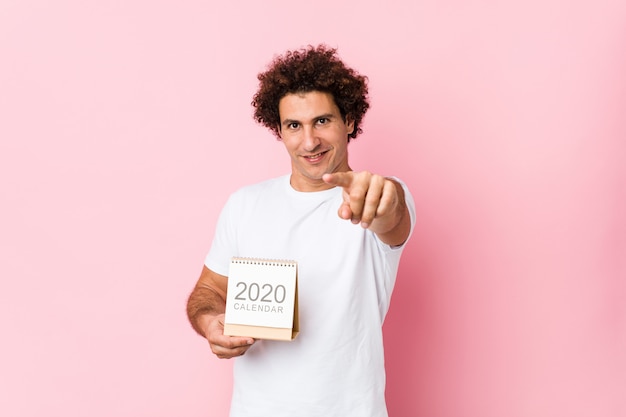 Jeune Homme Bouclé Caucasien Tenant Un Calendrier 2020 Sourires Joyeux Pointant Vers L'avant.