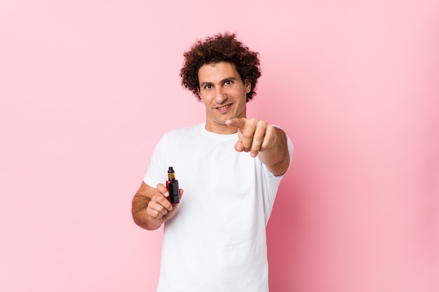 Jeune homme bouclé caucasien tenant une vape sourires gais pointant vers l'avant.