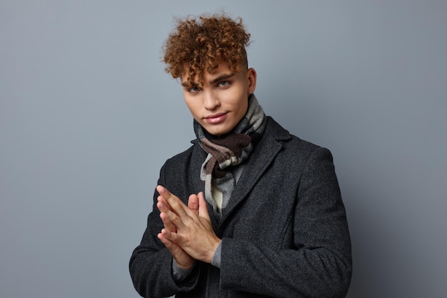 Jeune homme bouclé dans un manteau en studio posant sur fond gris