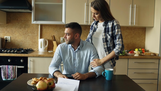 Jeune homme bouleversé lisant des factures impayées et étreint par sa femme le soutenant dans la cuisine à la maison