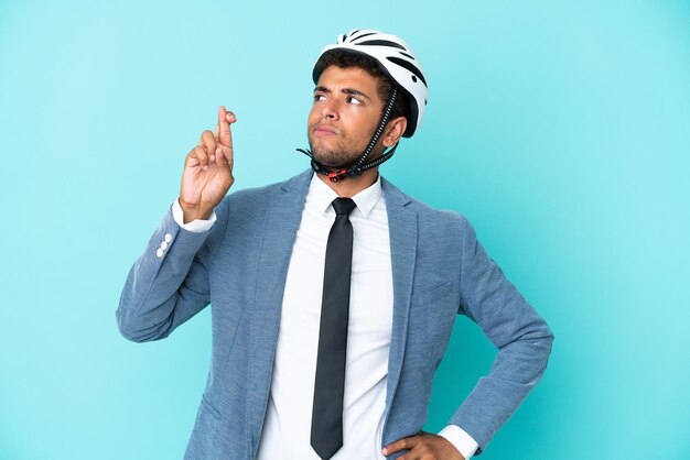 Jeune homme brésilien d'affaires avec casque de vélo isolé sur fond bleu avec les doigts qui se croisent et qui souhaitent le meilleur