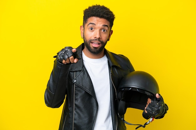 Jeune homme brésilien avec un casque de moto isolé sur fond jaune surpris et pointant vers l'avant