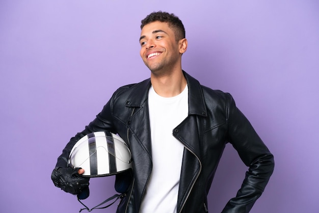 Jeune homme brésilien avec un casque de moto isolé sur fond violet posant avec les bras à la hanche et souriant