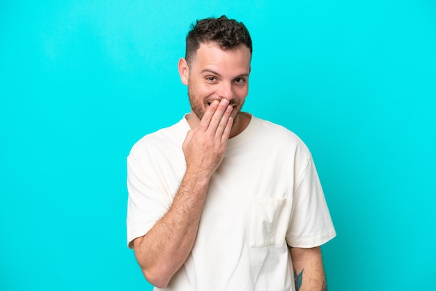 Jeune homme brésilien isolé sur fond bleu heureux et souriant couvrant la bouche avec la main