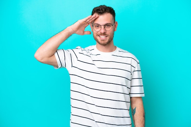 Jeune homme brésilien isolé sur fond bleu saluant avec la main avec une expression heureuse