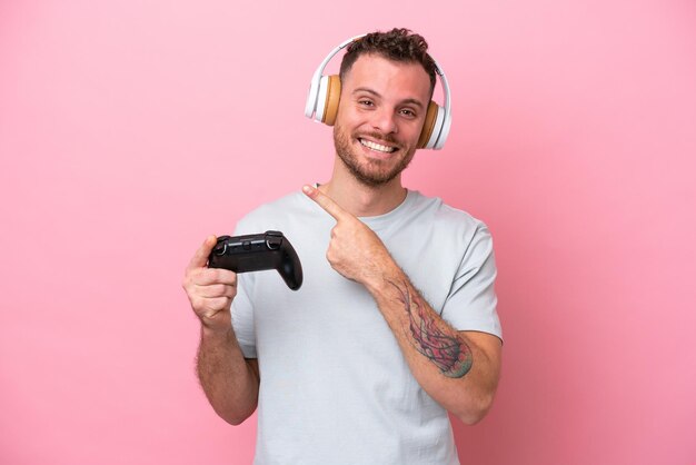 Jeune homme brésilien jouant avec un contrôleur de jeu vidéo isolé sur fond rose pointant vers le côté pour présenter un produit