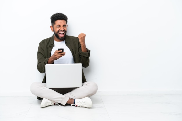 Jeune homme brésilien avec un ordinateur portable assis sur le sol isolé sur blanc avec téléphone en position de victoire