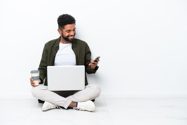 Jeune homme brésilien avec un ordinateur portable assis sur le sol isolé sur blanc tenant du café à emporter et un mobile