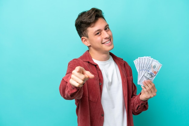 Jeune homme brésilien prenant beaucoup d'argent isolé sur fond bleu pointant vers l'avant avec une expression heureuse