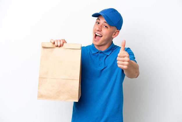 Jeune homme brésilien prenant un sac de plats à emporter isolé sur fond blanc avec les pouces vers le haut parce que quelque chose de bien s'est produit