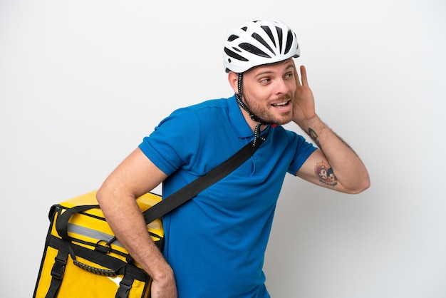 Jeune homme brésilien avec sac à dos thermique isolé sur fond blanc écoutant quelque chose en mettant la main sur l'oreille