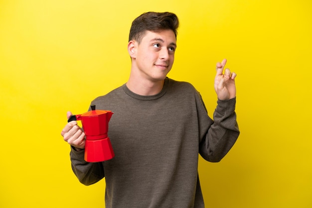 Jeune homme brésilien tenant une cafetière isolée sur fond jaune avec les doigts croisés et souhaitant le meilleur