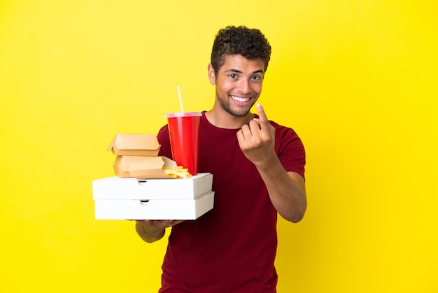 Jeune homme brésilien tenant des pizzas et des hamburgers fond isolé faisant un geste à venir