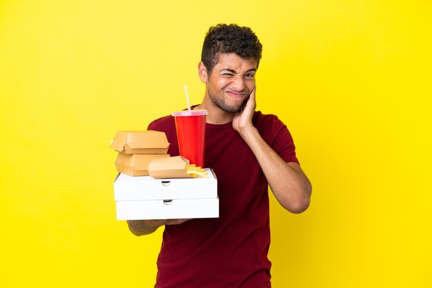 Jeune homme brésilien tenant des pizzas et des hamburgers fond isolé frustré et couvrant les oreilles