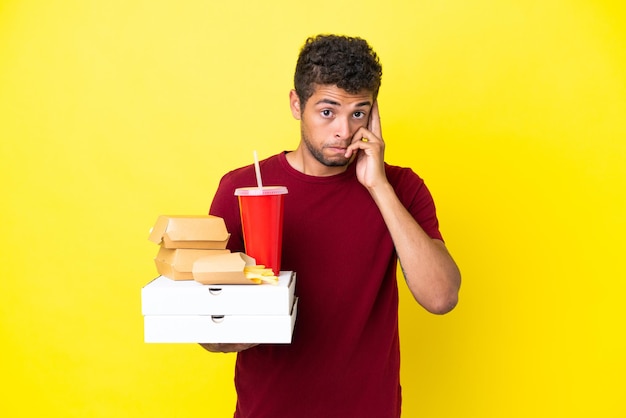 Jeune homme brésilien tenant des pizzas et des hamburgers fond isolé en pensant à une idée