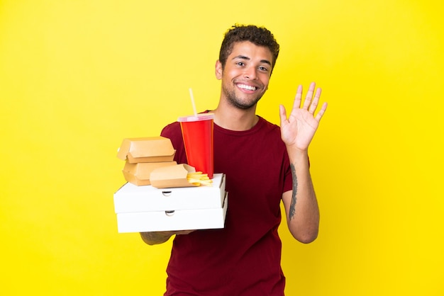 Jeune homme brésilien tenant des pizzas et des hamburgers fond isolé saluant avec la main avec une expression heureuse