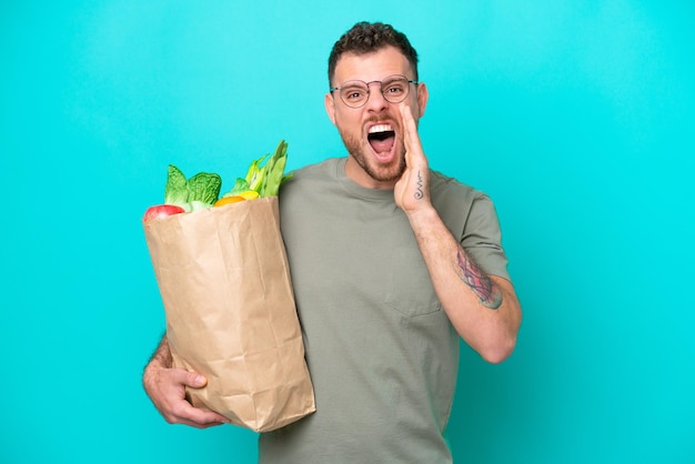 Jeune homme brésilien tenant un sac d'épicerie isolé sur fond bleu criant avec la bouche grande ouverte