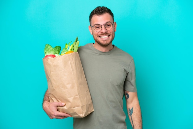 Jeune homme brésilien tenant un sac d'épicerie isolé sur fond bleu avec une expression faciale surprise