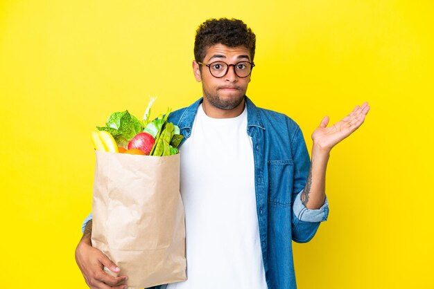 Jeune homme brésilien tenant un sac d'épicerie isolé sur fond jaune ayant des doutes tout en levant les mains