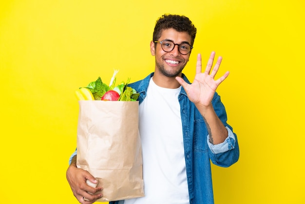 Jeune homme brésilien tenant un sac d'épicerie isolé sur fond jaune comptant cinq avec les doigts