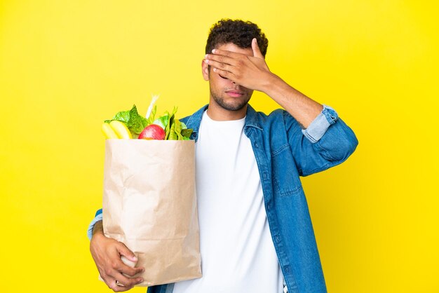 Jeune homme brésilien tenant un sac d'épicerie isolé sur fond jaune couvrant les yeux à la main. Je ne veux pas voir quelque chose