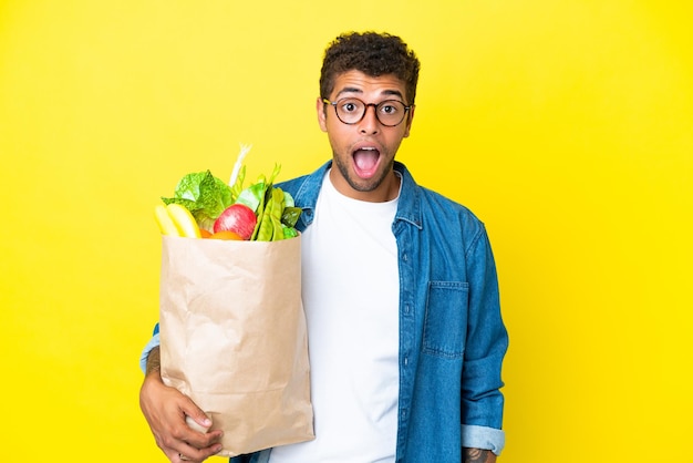 Jeune homme brésilien tenant un sac d'épicerie isolé sur fond jaune avec une expression faciale surprise
