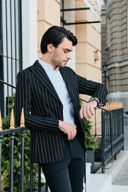 Jeune homme brune barbu attrayant en chemise blanche et veste rayée classique regardant rêveusement sur la montre-bracelet tout en se tenant sur la rue de la ville