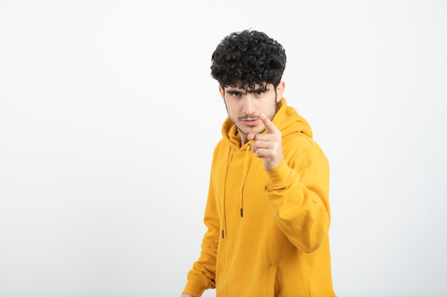 jeune homme brune en sweat à capuche jaune debout et pointant la caméra.