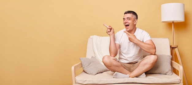 Jeune homme brunette excité en train de rire portant un t-shirt blanc décontracté assis sur le canapé contre le mur beige pointant vers la zone de publicité espace de copie pour le texte promotionnel