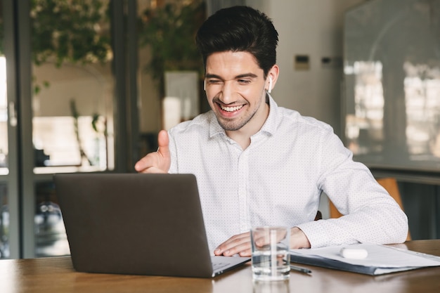 Jeune homme de bureau portant une chemise blanche et des écouteurs sans fil souriant alors qu'il était assis à table au bureau, et pointant le doigt sur l'écran de l'ordinateur portable pendant un appel vidéo ou un chat