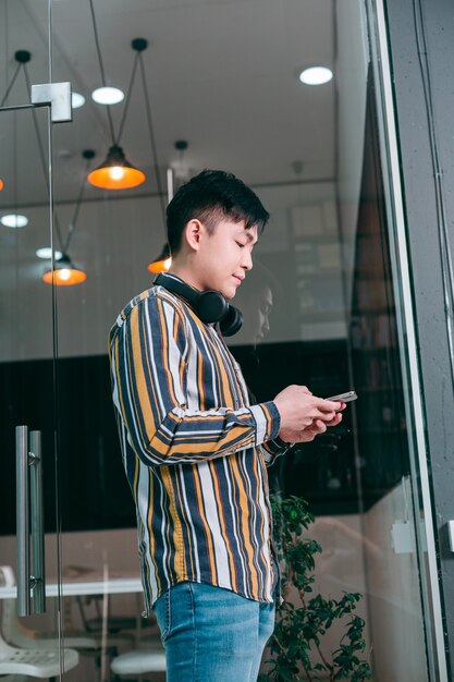 Jeune homme calme avec des écouteurs sur le cou, debout près de la porte vitrée et utilisant son smartphone