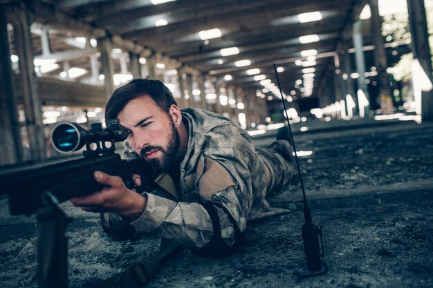 Un jeune homme calme et paisible est allongé sur le sol et vise. Il a l'air très sérieux. Guy utilise un fusil pour ça. Il regarde aussi à travers l'objectif. Guy attend.