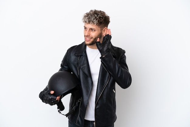 Jeune homme avec un casque de moto isolé sur fond blanc écoutant quelque chose en mettant la main sur l'oreille