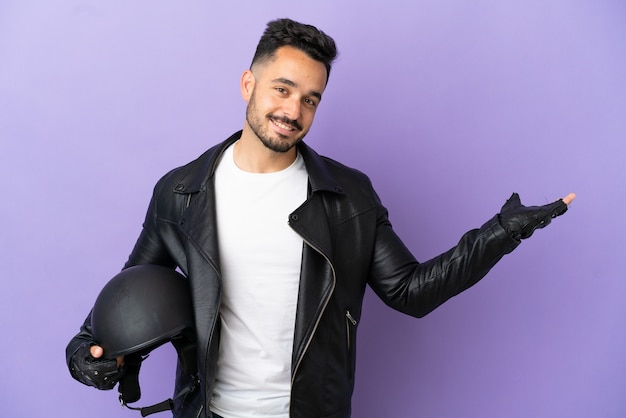 Jeune homme avec un casque de moto isolé sur fond violet tendant les mains sur le côté pour inviter à venir