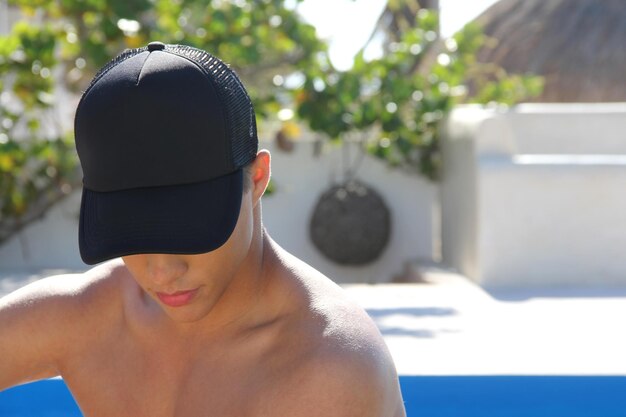 Jeune homme avec une casquette noire sur un fond de piscine. Notion d'été. Maquette de casquette de baseball vide.