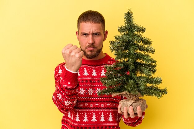 Jeune homme caucasien achetant un petit arbre pour Noël isolé sur fond jaune montrant le poing à la caméra, expression faciale agressive.