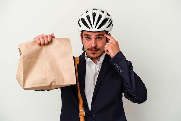 Jeune homme caucasien d'affaires portant un casque de vélo et tenant de la nourriture à emporter isolé sur fond blanc pointant le temple avec le doigt, pensant, concentré sur une tâche.