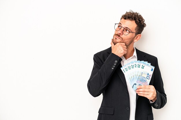 Jeune homme caucasien d'affaires tenant des billets isolés sur fond blanc regardant de côté avec une expression douteuse et sceptique.