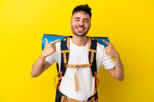 Jeune homme caucasien d'alpiniste avec un grand sac à dos d'isolement sur le fond jaune donnant un geste de pouce en l'air