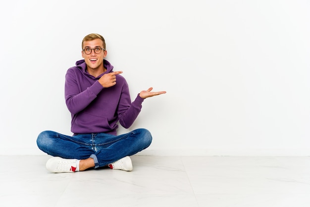 Jeune homme caucasien assis sur le sol excité tenant un espace de copie sur la paume.