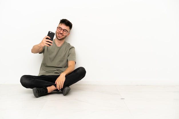 Jeune homme caucasien assis sur le sol isolé sur fond blanc faisant un selfie