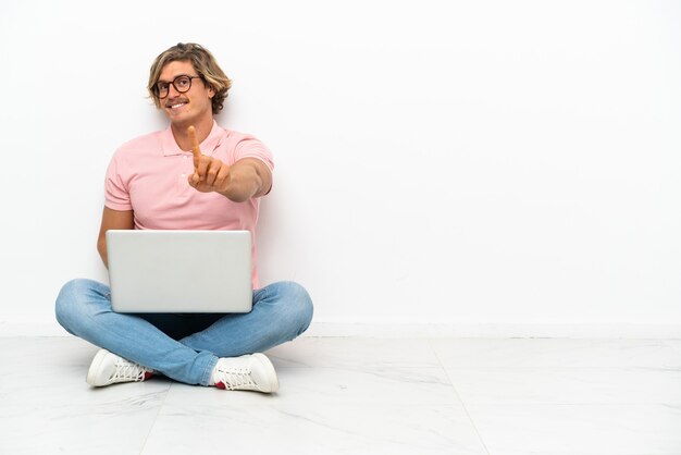 Jeune homme caucasien assis sur le sol avec son ordinateur portable isolé sur blanc montrant et soulevant un doigt