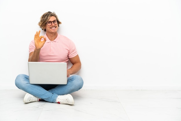 Jeune homme caucasien assis sur le sol avec son ordinateur portable isolé sur fond blanc montrant signe ok avec les doigts