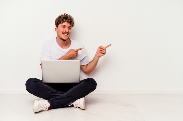 Jeune homme caucasien assis sur le sol tenant un ordinateur portable isolé sur fond blanc excité pointant avec l'index loin.