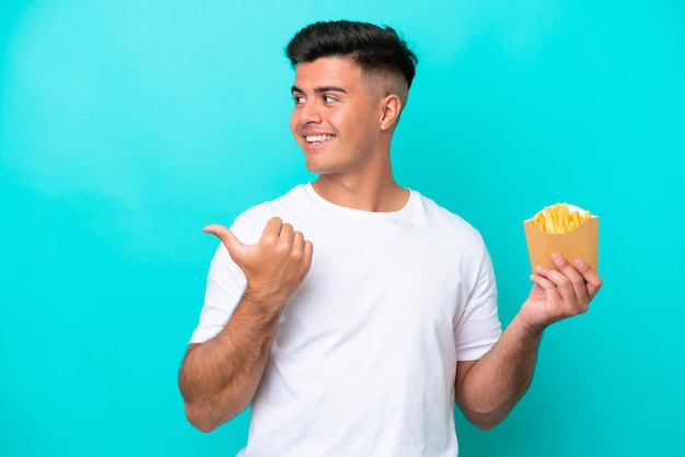 Jeune homme caucasien attrapant des frites isolées sur fond bleu pointant vers le côté pour présenter un produit