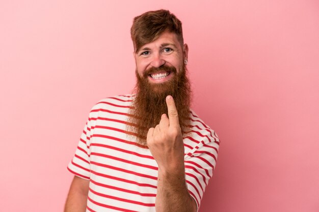 Jeune homme caucasien au gingembre avec une longue barbe isolé sur fond rose pointant le doigt vers vous comme s'il vous invitait à vous rapprocher.