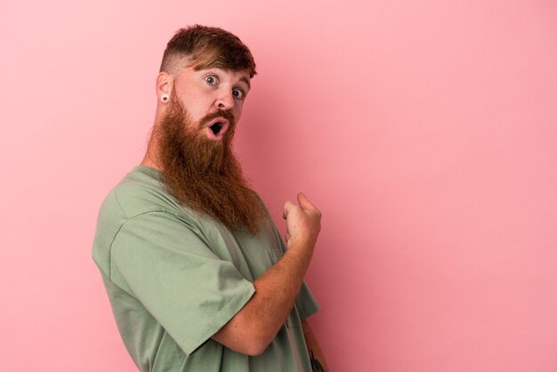 Jeune homme caucasien au gingembre avec une longue barbe isolé sur fond rose souriant et pointant de côté, montrant quelque chose dans un espace vide.