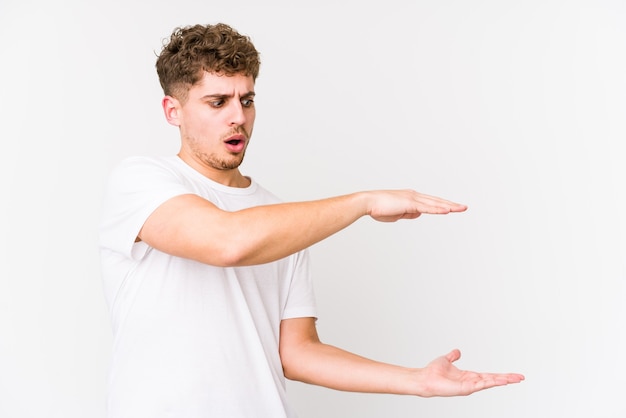 Jeune homme caucasien blond cheveux bouclés isolé choqué et étonné tenant un espace de copie entre les mains.