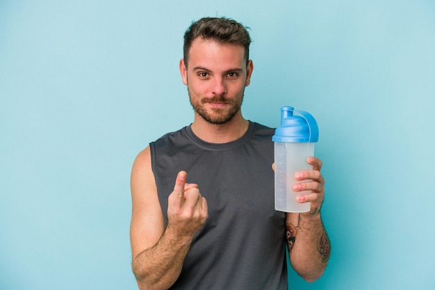 Jeune homme caucasien buvant un milkshake isolé sur fond bleu pointant du doigt vers vous comme s'il vous invitait à vous rapprocher